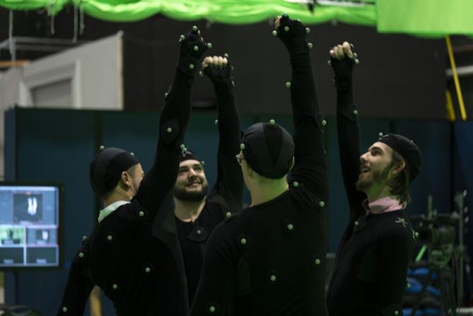 Quatre personnes vêtues d'un costume noir et d'un chapeau recouverts de petites boules vertes se tiennent en cercle, chacune d'entre elles levant un bras