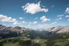 Rocky mountains and lush green fields under a sunny sky dotted with white clouds