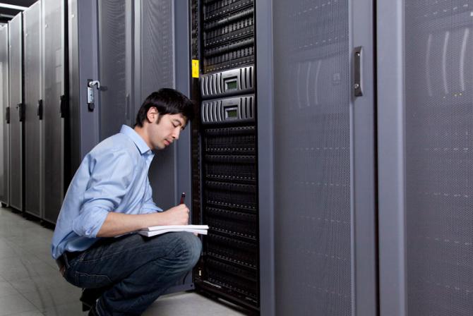 A person in a server room crouches next to an open server door and write data in a notebook resting on his knee..