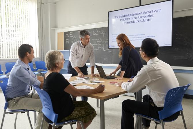 Five people work together at a conference table, with a blackboard and white screen in the background