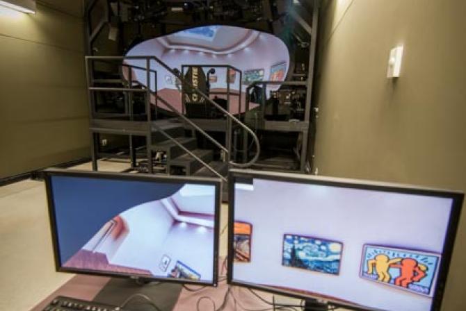 Two workstations sit on a table in the forefront, a set of stairs at the back of the room lead to a large apparatus displaying a room on a large oval shaped screen. Portions of the apparatus' displays appear on the monitors.
