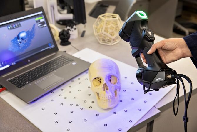 A hand holds a laser scanner over a skull on a table.