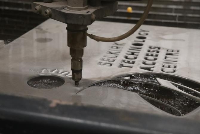 A waterjet cuts out shapes and letters in a sheet of metal.