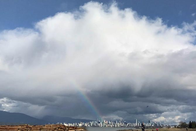 A rainbow shines down from a massive cloud looming over a city.