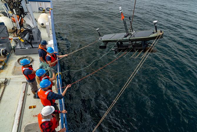 People wearing orange vests and hard hats line the side of a deck as they hold ropes connected a large piece of equipment floating in the water below.
