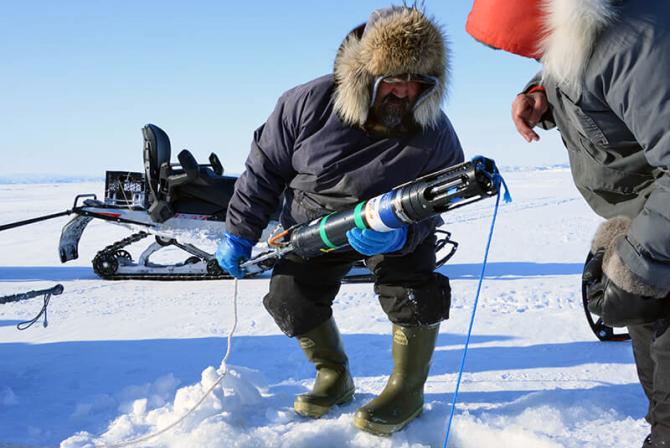 Two people wearing winter clothing stand on the ice as they examine a large sensor pulled from the water.