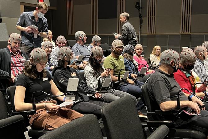 A group of people seated in an auditorium wear EEG caps connected to their armrests.