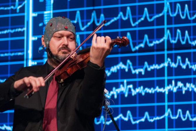 Un musicien portant un électro-cap joue du violon devant un grand écran affichant des lignes ondulées.