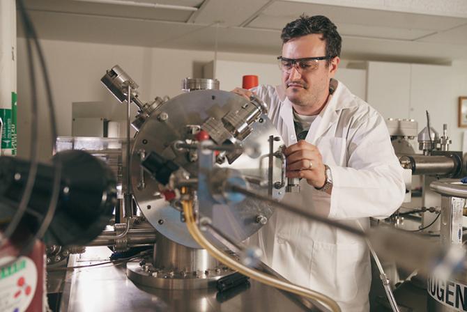 Researcher wearing a white lab coat and safety glasses looking at a sample of aluminum on the sample holder inside the GD-MS.